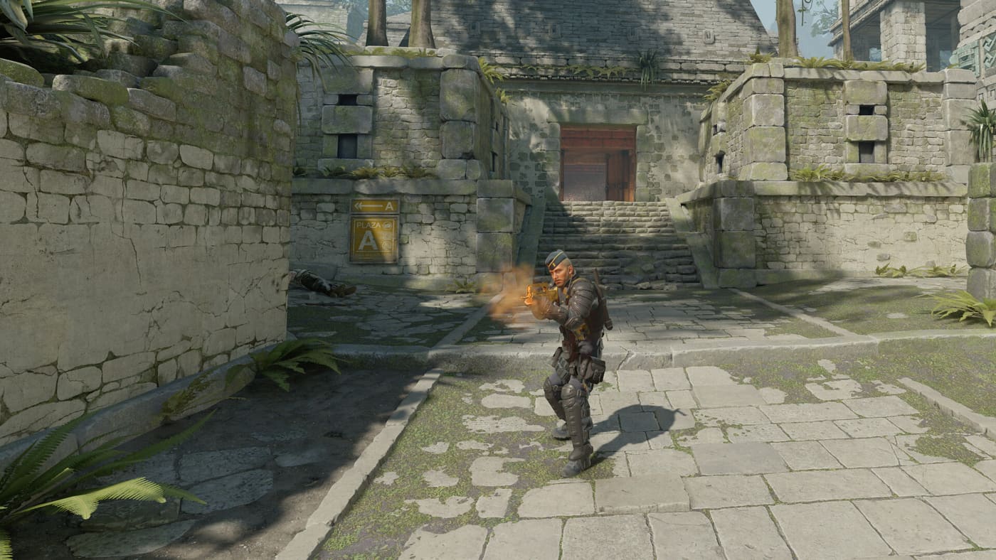 A Counter-Strike 2 player fires in a sunlit stone courtyard by an A site marker, muzzle flash bright and a body already down on the ground to his left, seen from behind in the third-person view a demo camera gives you. Official Counter-Strike 2 screenshot supplied by Valve.